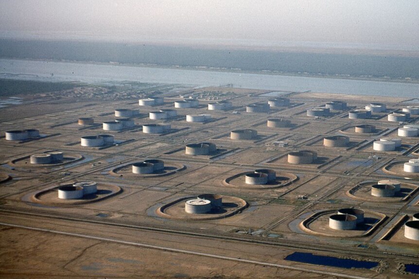 Aerial view of long rows of petroleium storage containers in desert setting beside a canal.