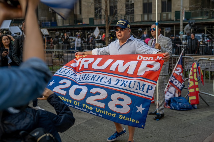 A man holds a flag that says 'Trump Keep America Trump 2028' in front of metal barricades outside a courthouse.
