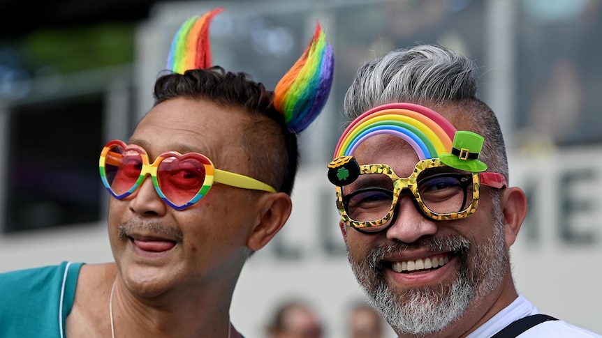 two males wearing rainbow coloured glasses and headgear sitting down at a stadium
