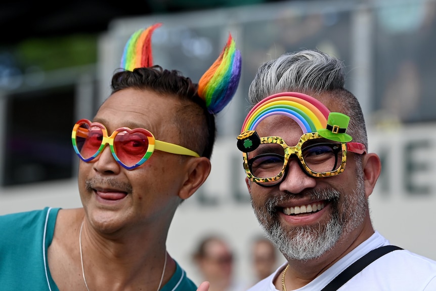 two males wearing rainbow coloured glasses and headgear sitting down at a stadium