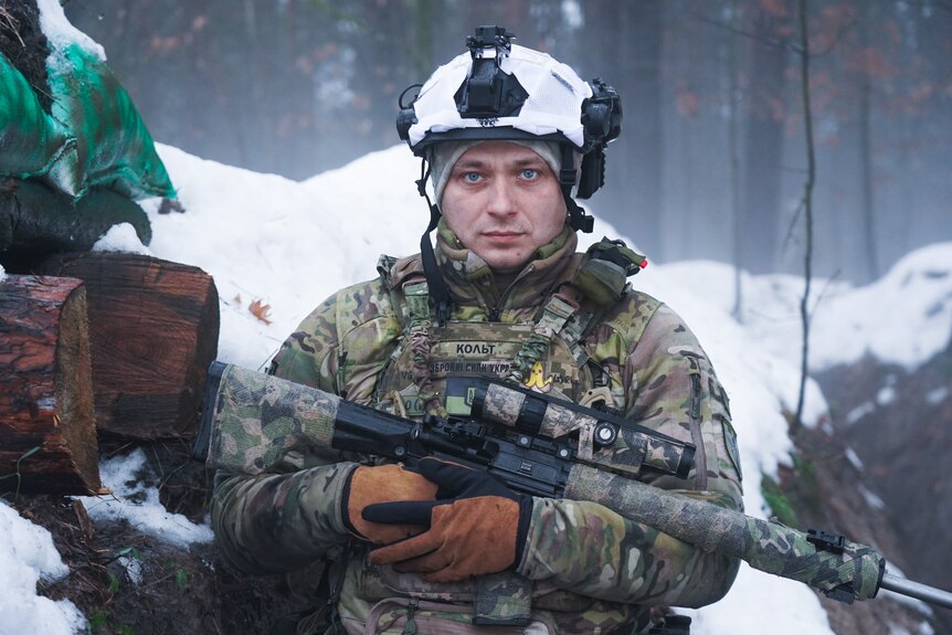 A man wearing army fatigues and holding a gun wears a snow covered helmet in a field.