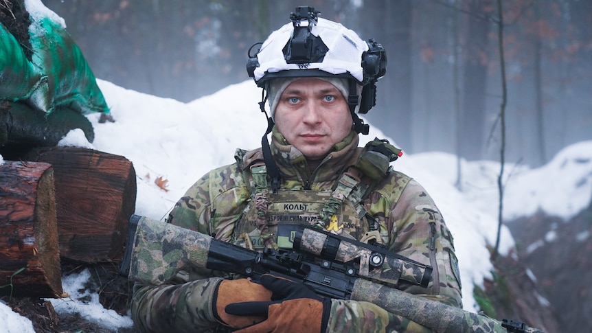 A man wearing army fatigues and holding a gun wears a snow covered helmet in a field.