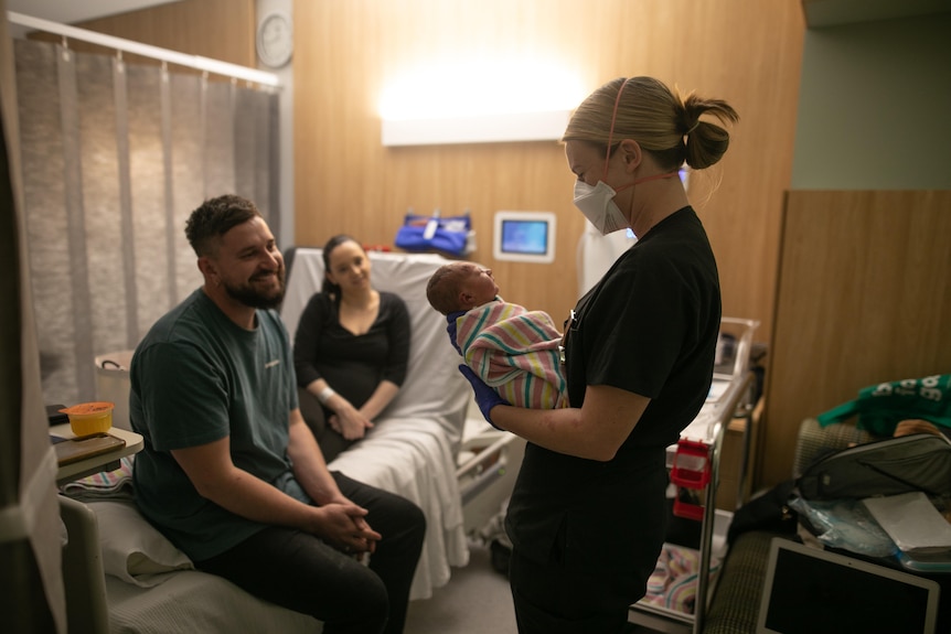 Taryn and Nathan sit on a hospital bed smiling, as a midwife holds their newborn baby.