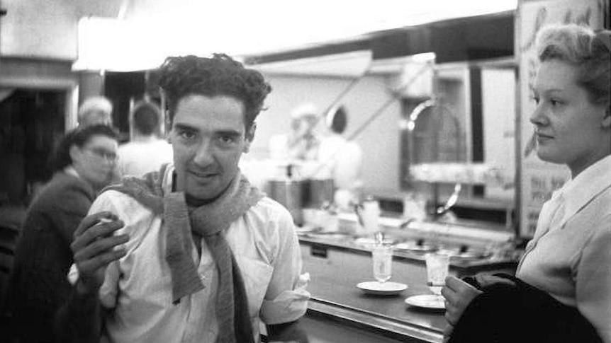 Black and white photo of a man and woman at a bar. Man looks at camera, woman looks at man. 