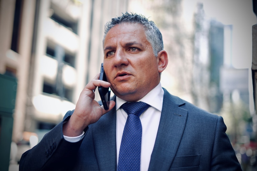 A man wearing a suit and tie holds a phone to his right ear.