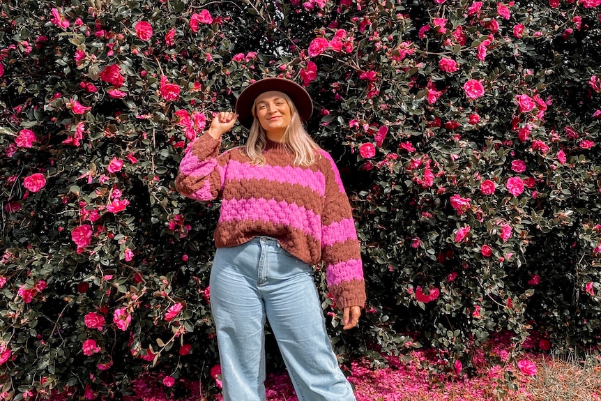 A young woman in a pink and red striped jumper, jeans and a hat poses smiling in front of a large bush with pink flowers.