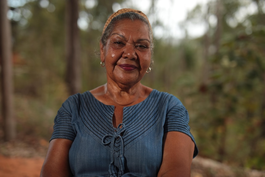 A woman wearing a headband and blue dress looks at the camera and is surrounded by trees.