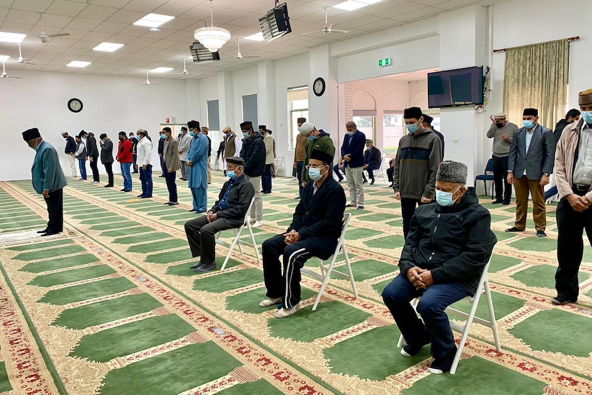Men stand and sit on chairs praying