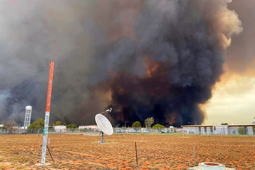 Large plumes of black smoke erupt behind a fenced off military base