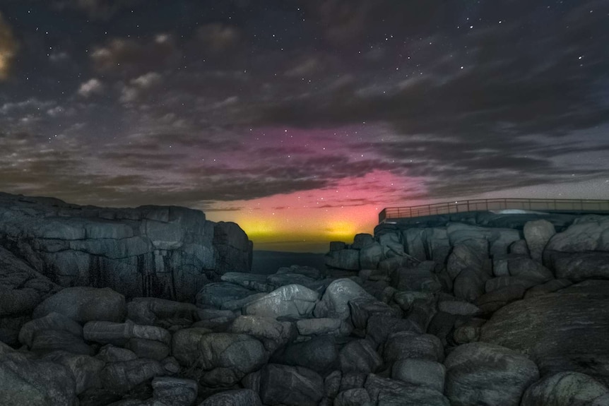 The night sky glows pink and yellow over a dark, rocky shore.