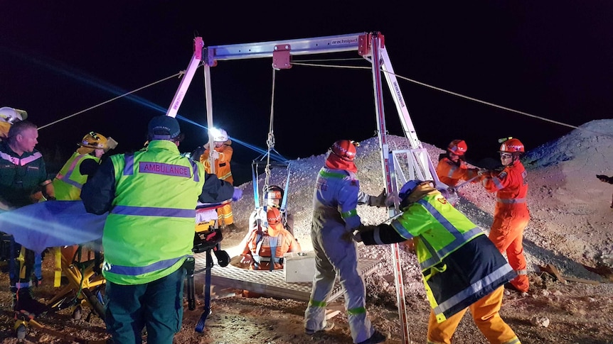 A dozen rescuers and a winch at the top of a mine shaft lit up at night time.