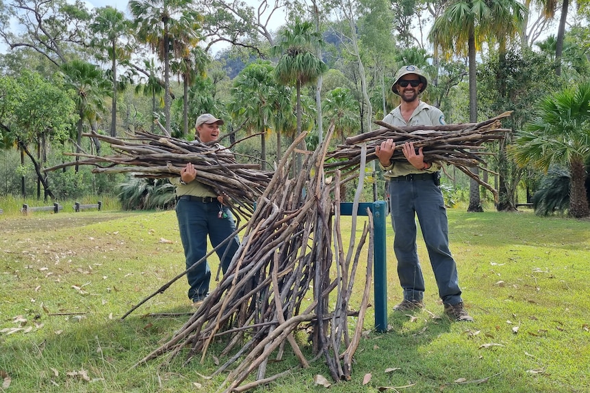 Two rangers holding large bundles of sticks in front of a large vertical pile of sticks