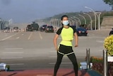 A woman in a face mask, leggings and t-shirt dancing with military vehicles behind her