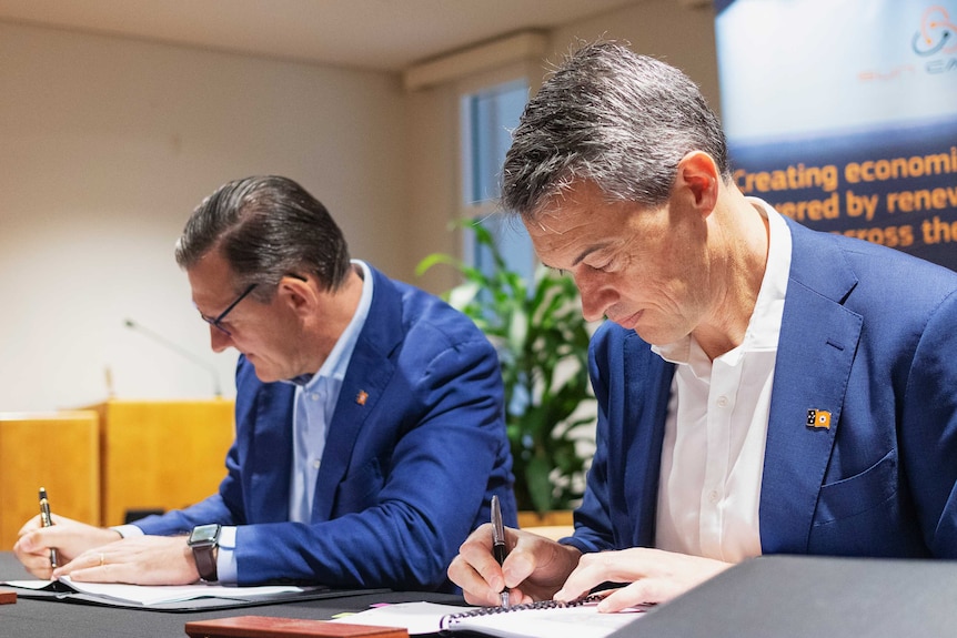 Two men in blue suits sit side by side signing forms and smiling.