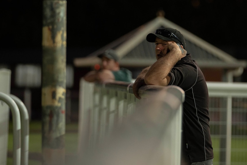 A man talks on the phone while leaning on a fence at a racecourse.