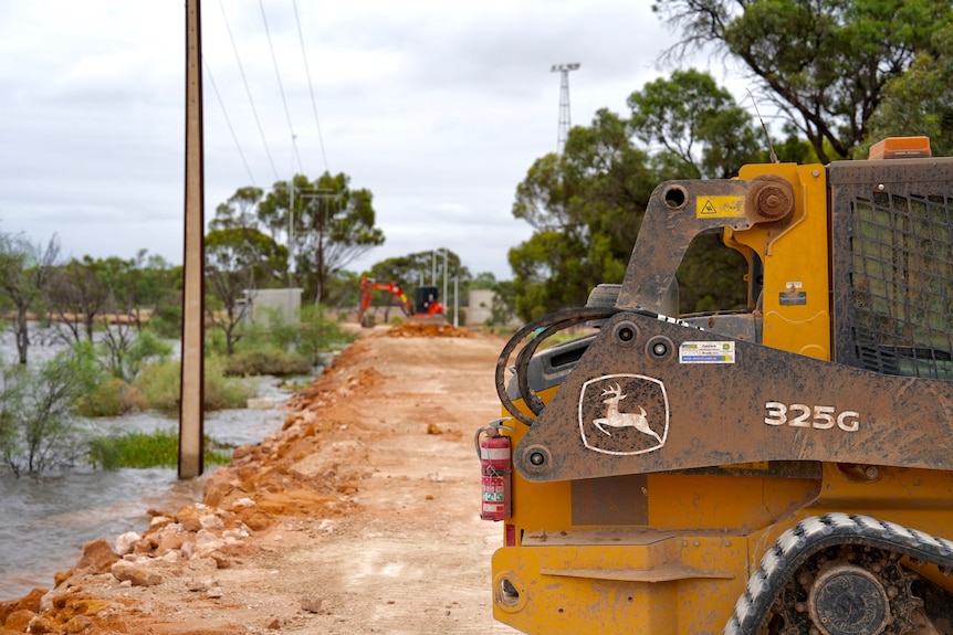 A side view of an excavator, on top of a flood levee with brown rocks and clay. It's next to a blue river, the sky is cloudy.