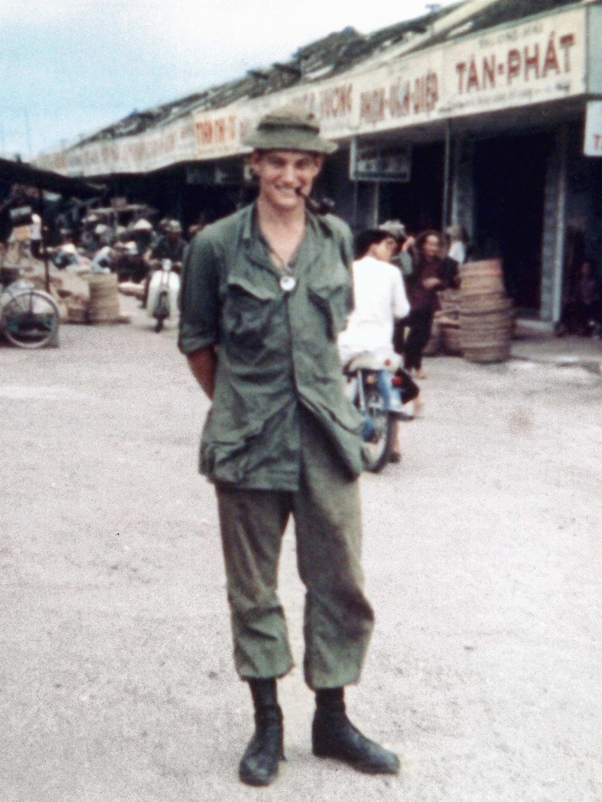 A man wearing khaki with a pipe in his mouth smiles at the camera.