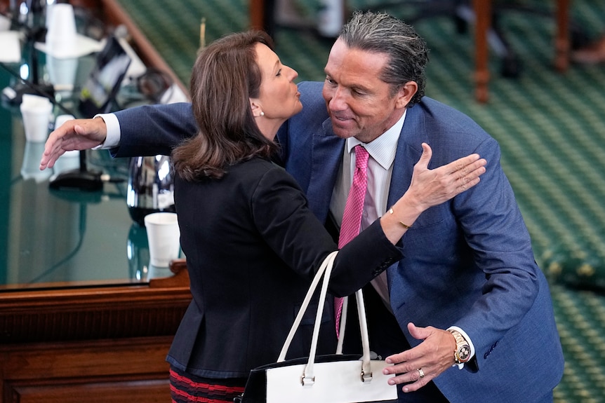 A man in a suit and tie hugs and kisses a woman in a suit who is holding a handbag, inside a legislative building.
