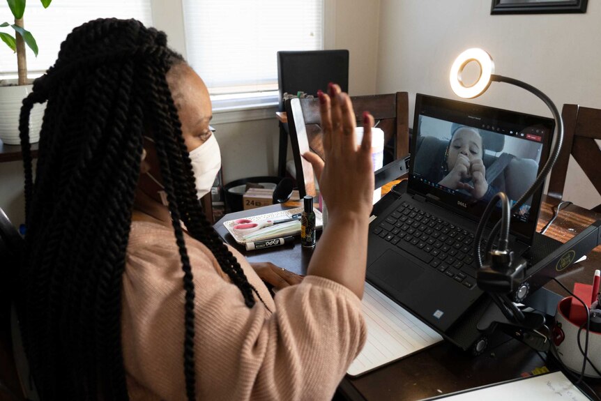 An African American woman with box braids and a face mask gestures to a child on video call