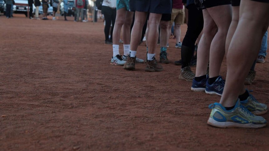 Shoes, socks and legs of potential Uluru climbers stand on red dirt.