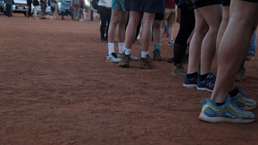 Shoes, socks and legs of potential Uluru climbers stand on red dirt.