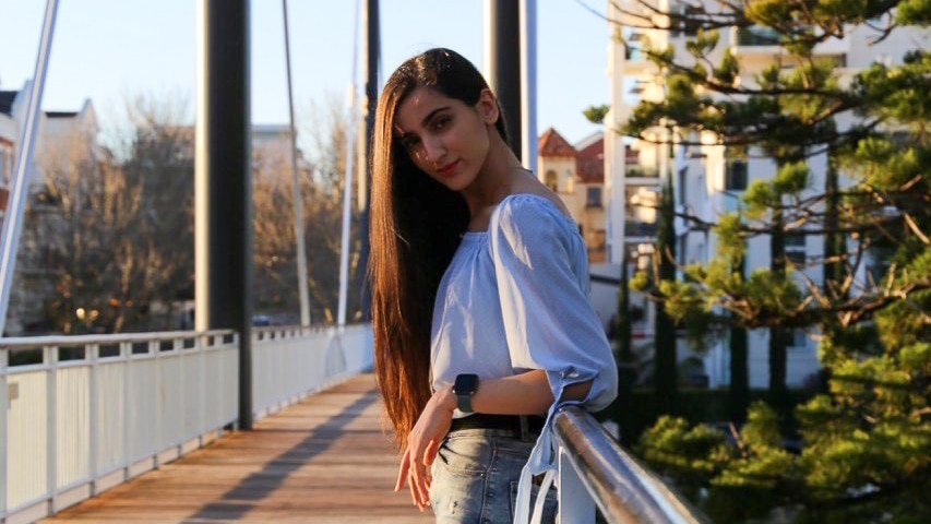Young woman with long hair and white top and jeans stands on bridge.