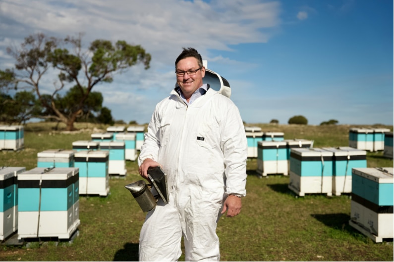 A man standing between boxes of beehives in a beekeeping suit, smiles to camera