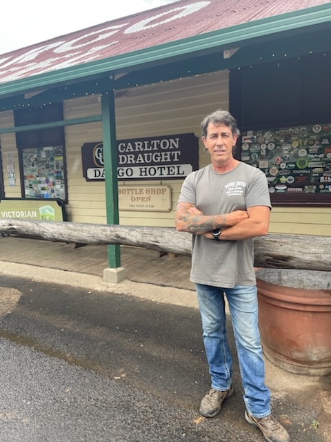 Una foto de un hombre con una camiseta gris y un tatuaje parado frente a un edificio llamado Hotel Dargo.