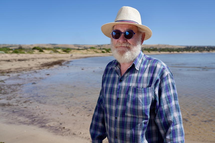 A man wearing a long sleeve shirt, wide brimmed hat and sunglasses stands on a beach