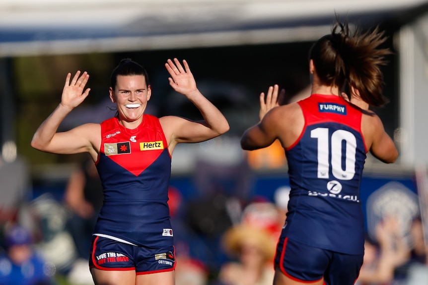 A smiling Melbourne AFLW player comes forward to high-five a teammate after kicking a goal.