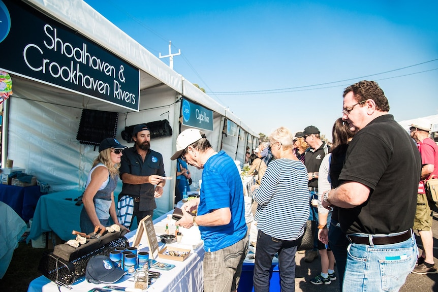 A crowd of people gathered near outdoor stalls including one with the sign Shoalhaven and Crookhaven Rivers.