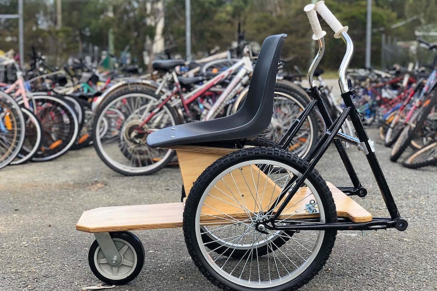 A wheelchair made from old bike parts in front of a collection of old bikes