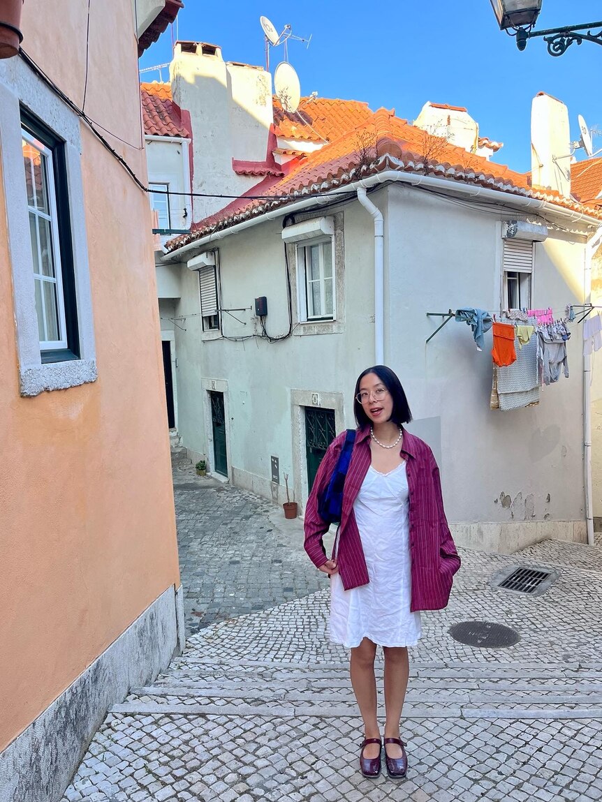 Writer Maggie Zhou on a quiet street in Porto, Portugal wearing a white dress with a button-up shirt layered on top.
