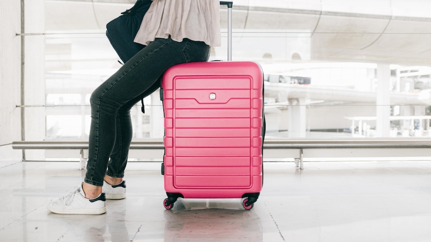 A person wearing dark jeans and runners sits on a bright pink suitcase while holding a phone in an empty airport.