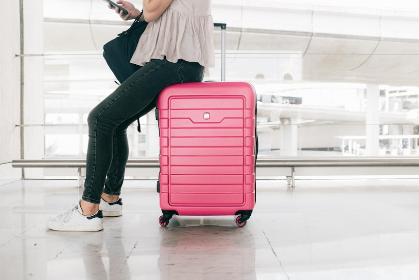 A person wearing dark jeans and runners sits on a bright pink suitcase while holding a phone in an empty airport.