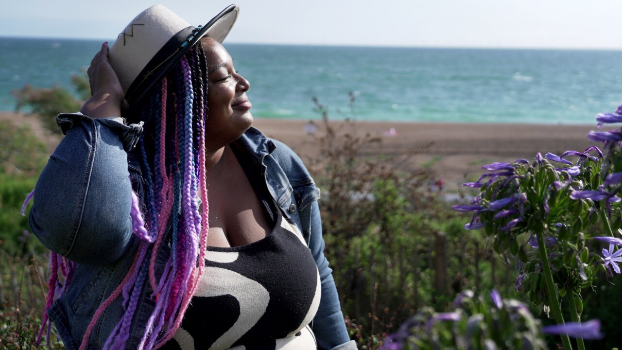 A woman with a beach behind her, turning her head with her eyes closed and smiling with the sun on her face