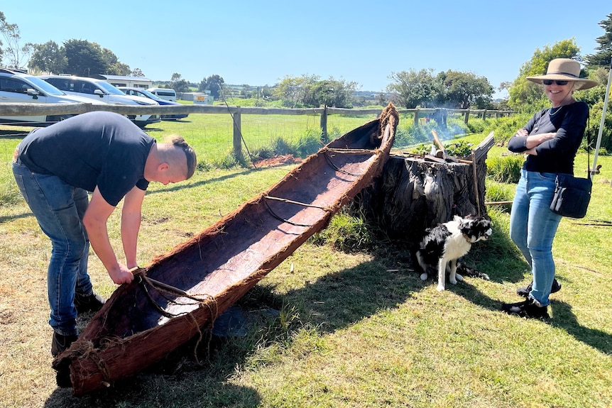 a man works on a large canoe, with a dog and a woman standing nearby.