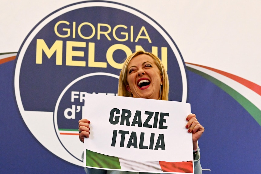 A middle-aged blonde woman smiles as she holds a sign in front of her saying "Grazie Italia"
