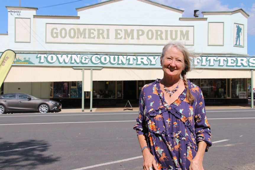 A woman in a purple floral dress smiles at the camera, a big shop called the 'Goomeri Emporium' across the road behind her