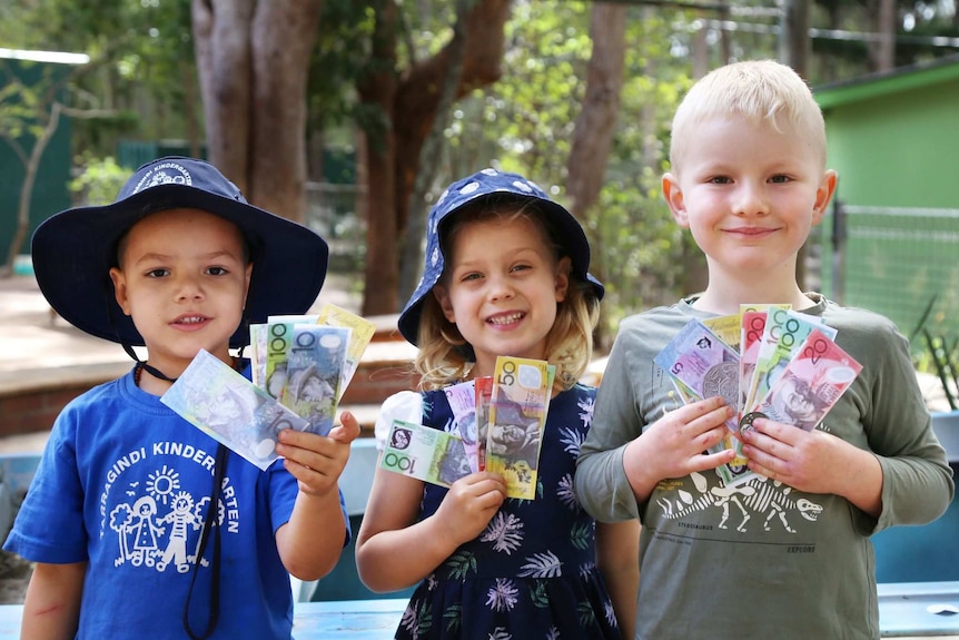 Three five-year-olds stand together in a kindy playground, holding play money and smiling.