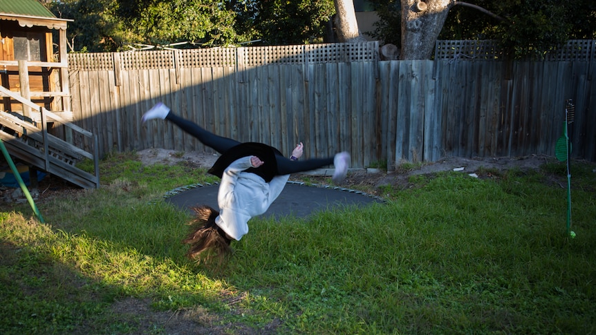 A girl does a somersault, legs split, in a grassy backyard, a cubby house in the rear.