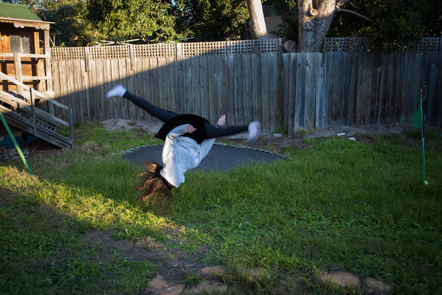 A girl does a somersault, legs split, in a grassy backyard, a cubby house in the rear.