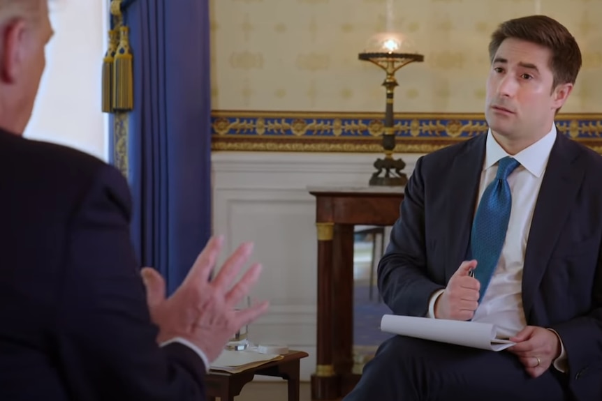 A man gestures while another listens with a notepad and pen.