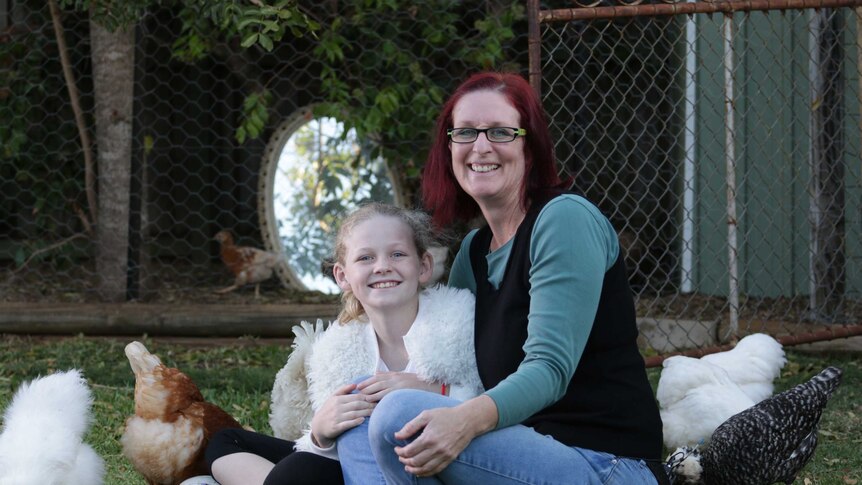 Little girl in a white fluffy jacket and her mum next to her sit in a yard among chickens