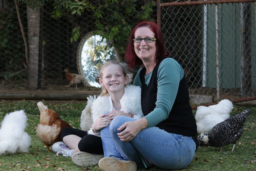 Little girl in a white fluffy jacket and her mum next to her sit in a yard among chickens