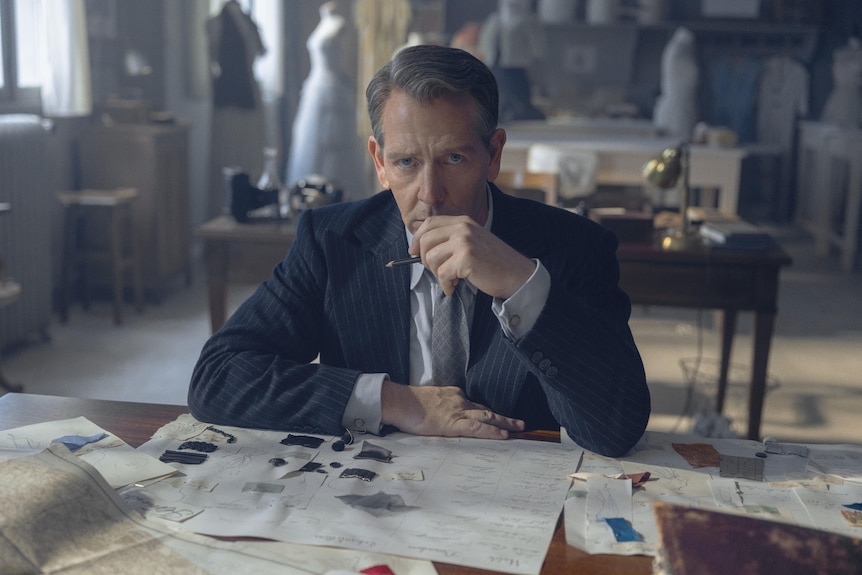 A concerned man sits at a table in a suit with a pen in hand, papers with fashion designs sprawled out in front of him.