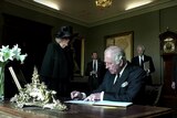 king charles sits at the table signing the book, camilla by his side and aides hastening towards him in the background