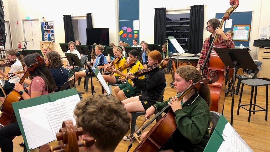 A wide shot of students in a rehearsal room holding string instruments and reading music. 
