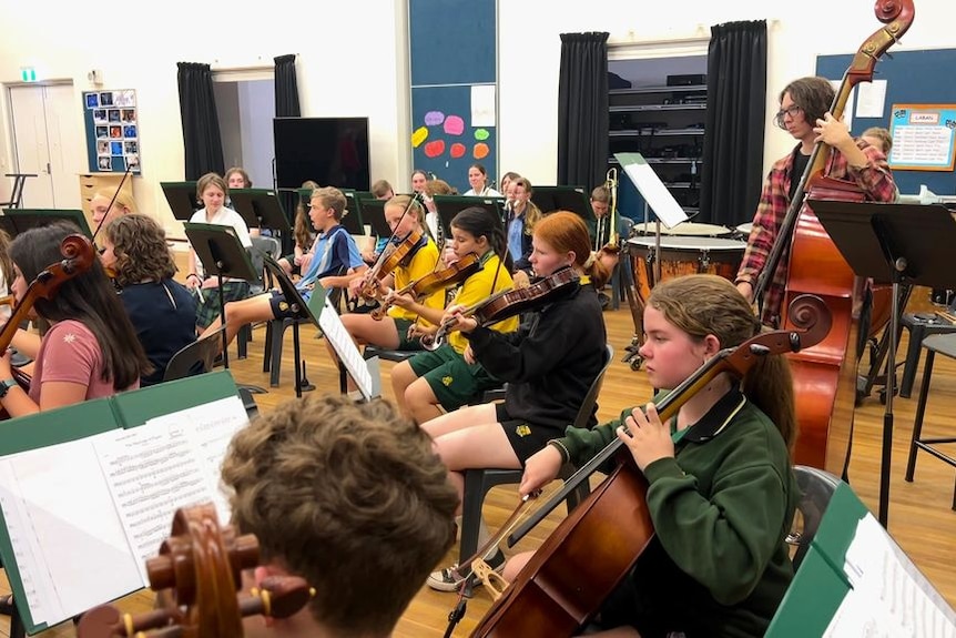 A wide shot of students in a rehearsal room holding string instruments and reading music. 