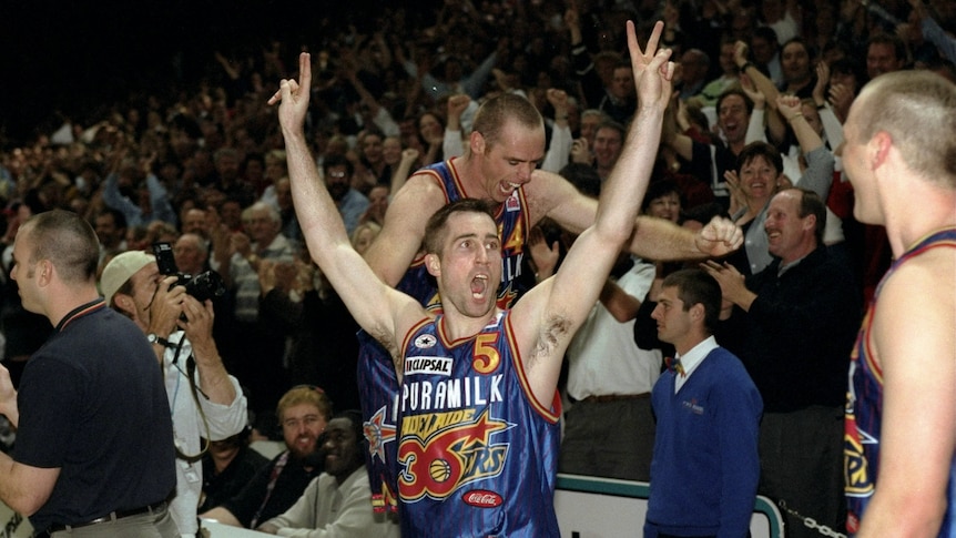 A basketball player with his hands up in a V pose while another man walks on seats among a cheering crowd